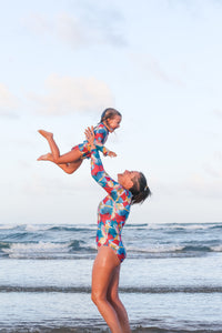 A woman throws a child joyfully in the air. They wear matching brightly patterned long sleeved UV protective handmade swimsuits made on the Sunshine Coast.